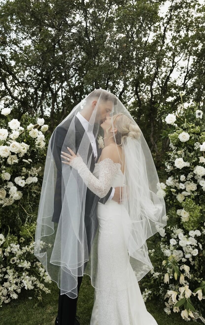 A bride and groom kissing beneath her veil between two arrangements of white flowers